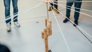 Holzobjekt hängt an Schnüren in einer Galerie, umgeben von Menschen.