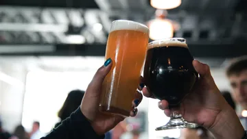 Two glasses toast, surrounded by people in a bar.