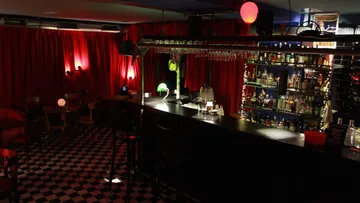 Rote Vorhänge umgeben eine beleuchtete Bar mit Flaschen.
