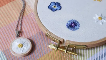 Embroidered flowers on fabric, handmade necklace next to it.