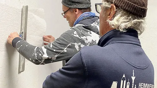 Two craftsmen measure and work on a wall.