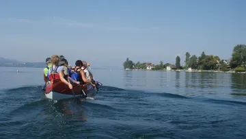 Ruderboot mit Personen fährt auf ruhigem Wasser, umgeben von Natur.
