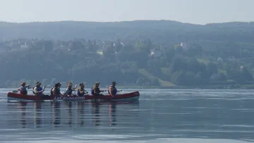 Kanu mit acht Personen paddelt ruhigen See vor hügeliger Landschaft.