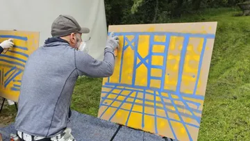A man is spraying blue patterns on a yellow canvas outside.
