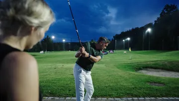 Man hits golf ball; woman watches on a golf course.