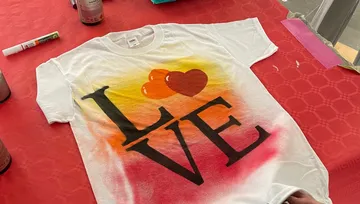 Weißes T-Shirt mit buntem `LOVE`-Design auf rotem Tisch.