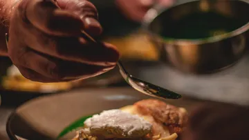 Eine Hand hält einen Löffel und gibt Essen auf einen Teller.