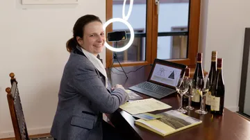 Frau sitzt lächelnd am Tisch mit Weinflaschen und Laptop.