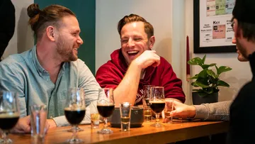 Zwei Männer lachen, Getränke auf einem Tisch, entspannte Atmosphäre.