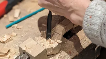 Eine Hand schnitzt Holz mit einem Werkzeug auf einer Werkbank.