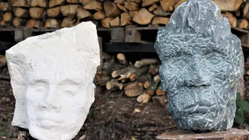Two facial sculptures stand in front of a pile of wood.