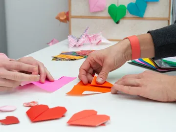 Zwei Hände falten Origami-Herzen aus buntem Papier am Tisch.