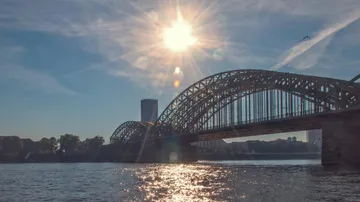Brücke reflektiert Sonnenlicht über ruhigem Wasser, Stadt dahinter.