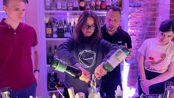 Person mixt Getränke an einer Bar, Gäste beobachten interessiert.