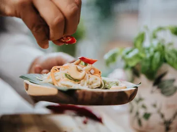 A hand sprinkles chilli on a pasta plate in a bright environment.