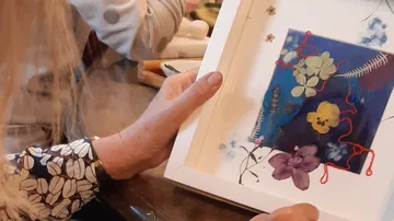 One hand holds a picture frame with pressed flowers.