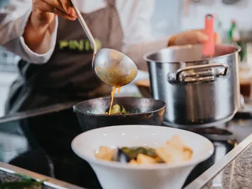 Cook pours soup into bowl surrounded by cooking utensils.