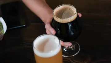 Schwarzes Bierglas in Hand; helles Bier daneben auf Tisch.