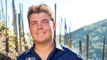 Mann mit Glas in Weinberg, Sonne scheint, Berge im Hintergrund.