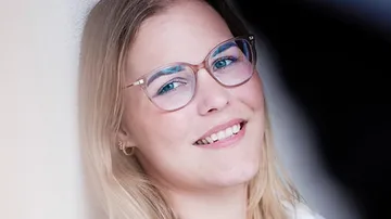 Junge Frau lächelt, steht an einer Wand, mit Brille.