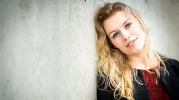 Junge Frau lächelt, lehnt an einer grauen Wand.