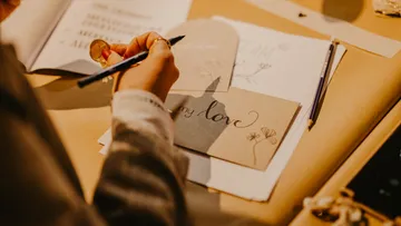 Eine Hand zeichnet auf eine Postkarte, umgeben von Papieren.