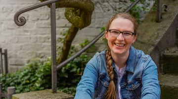 Young woman smiling, sitting on stones in front of stairs, surrounded by plants.
