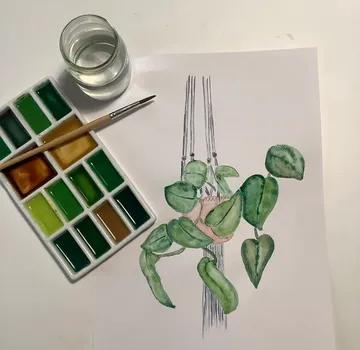 Topfpflanze hängt, Wasserfarben und Pinsel daneben, auf Tisch.