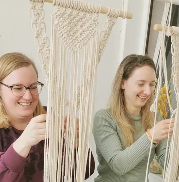 Two women weave macrame in a bright room.