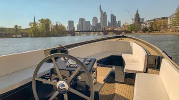 Boot mit Steuerrad fährt auf Fluss, skyline im Hintergrund.