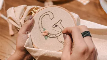 Eine Hand stickt ein Muster auf einem Stoffrahmen.