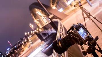 Camera on a tripod photographs an illuminated bridge at night.