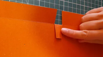 Orange paper is folded while one hand is holding it.