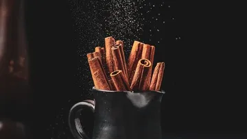 Zimtstangen stehen in einer schwarzen Tasse, es wird gestreut.
