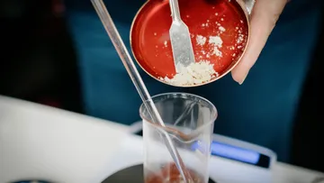 A hand holds a container and pours powder into a beaker.