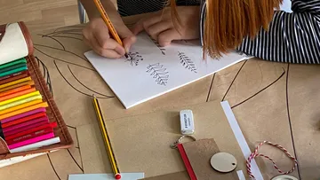 Person draws with pencil on paper surrounded by pens.
