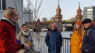 Group of people listen to a speaker in front of a bridge.