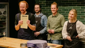 Four people are smiling behind a table in a kitchen.
