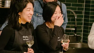 Two women laugh, holding wine glasses in a kitchen.