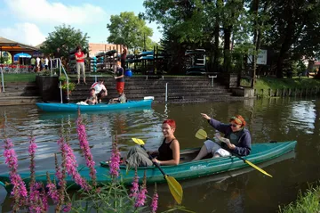 Two kayaks travel in water, with people and plants.