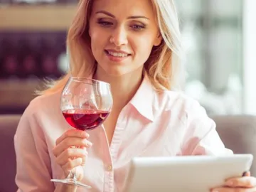 Woman holding wine glass, looking at tablet in cafe.