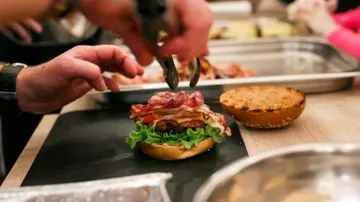 A hand covers a burger with bacon and salad.
