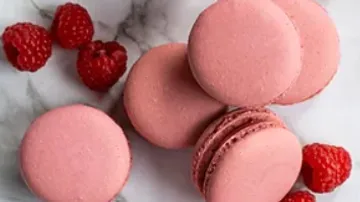 Pink macaroons lie on a marbled surface next to raspberries.
