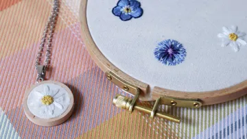 Kette mit gesticktem Blumenanhänger neben Stickrahmen auf Stoff.