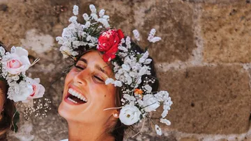 Lächelnde Frau mit Blumenkranz vor steinerner Wand.