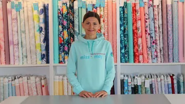 Woman in a fabric shop, stands in front of a wall with colorful rolls of fabric, smiles friendly at the camera.