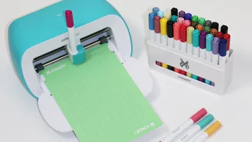 Kreativmaschine schneidet grünes Papier, umringt von Stiften.