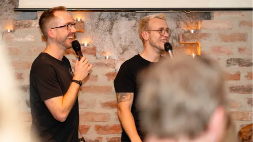 Two men laughing and holding microphones indoors.