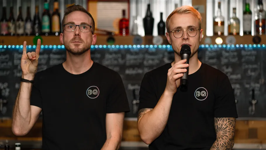 Two men in T-shirts stand in front of a bar, one is talking.