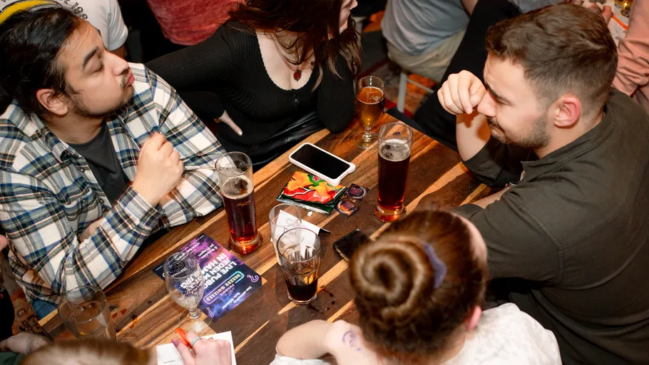 Eine Gruppe von Personen sitzt an einem Holztisch in einer Bar, trinkt Bier und unterhält sich angeregt, während Snacks und Getränke auf dem Tisch stehen.